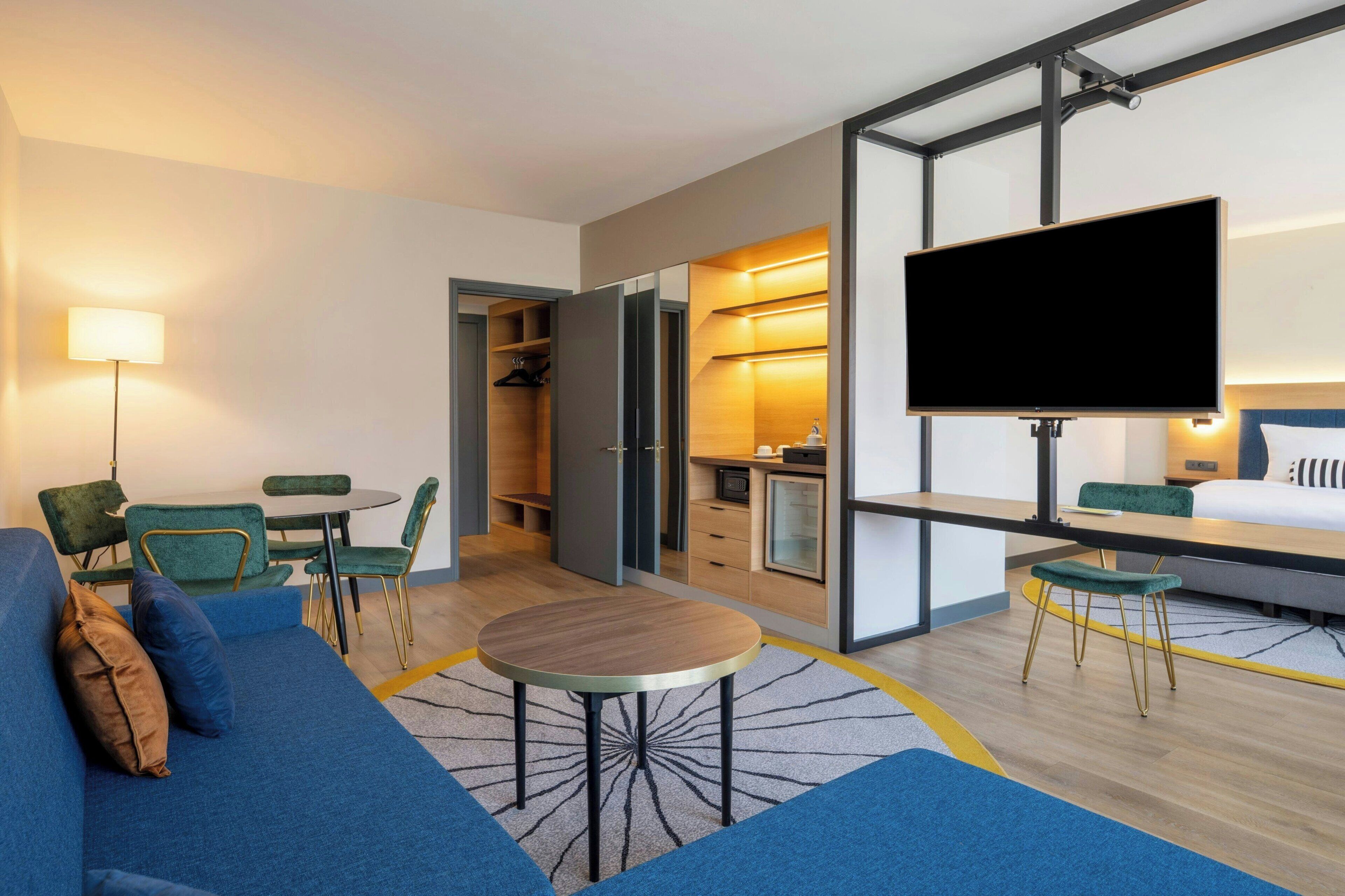 A modern hotel room with a sofa, a round table, a TV, a kitchenette, and a walk-in closet.