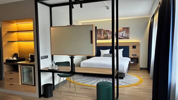 Ein Hotelzimmer mit einem Bett, einem Schreibtisch mit Computer, einem Stuhl, einem kleinen Tisch mit Tablett, einem Mülleimer, einem grünen Pouf und einem Bild an der Wand.