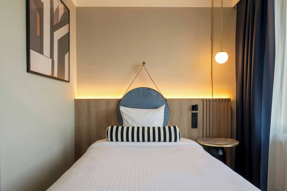 Una habitación de hotel con una cama, una lámpara redonda fijada a la pared, una pequeña mesa redonda y un panel de control en la pared.