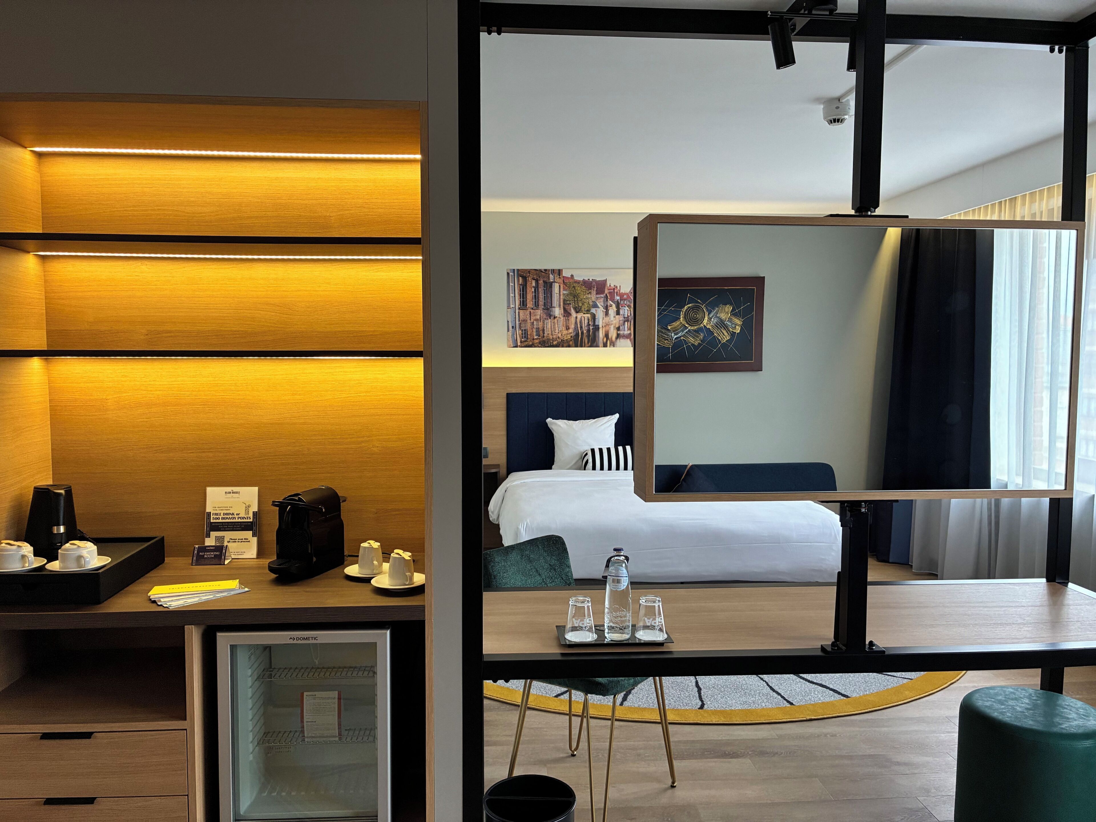 A hotel room with a bed, bedside table, mirror, and a view of a cityscape.