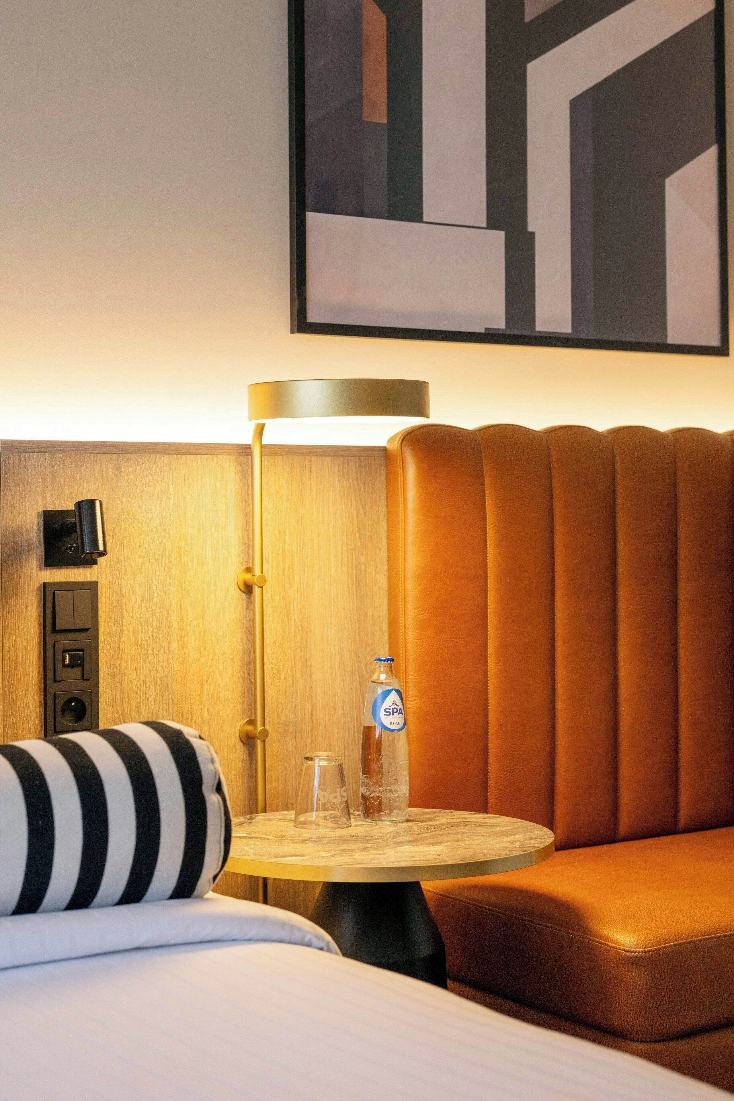 A hotel room with a bed, a round table with a bottle, a lamp, and a wall-mounted control panel.