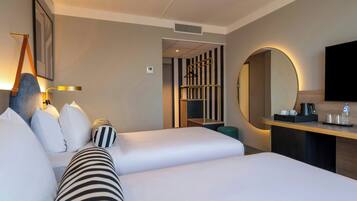 Une chambre d’hôtel avec deux lits, un bureau avec un miroir, une télévision et un coin salon comprenant une chaise et une petite table.