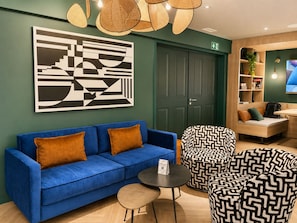 Un sofá azul con cojines marrones, un sillón estampado, una mesita redonda pequeña y una obra de arte abstracto colgada en la pared.
