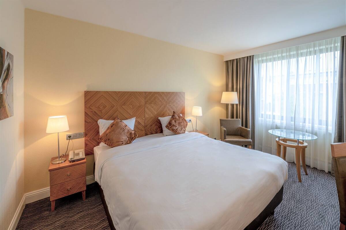Habitación de hotel con una cama grande, dos mesitas de noche con lámparas, una silla, una pequeña mesa y una ventana con cortinas.