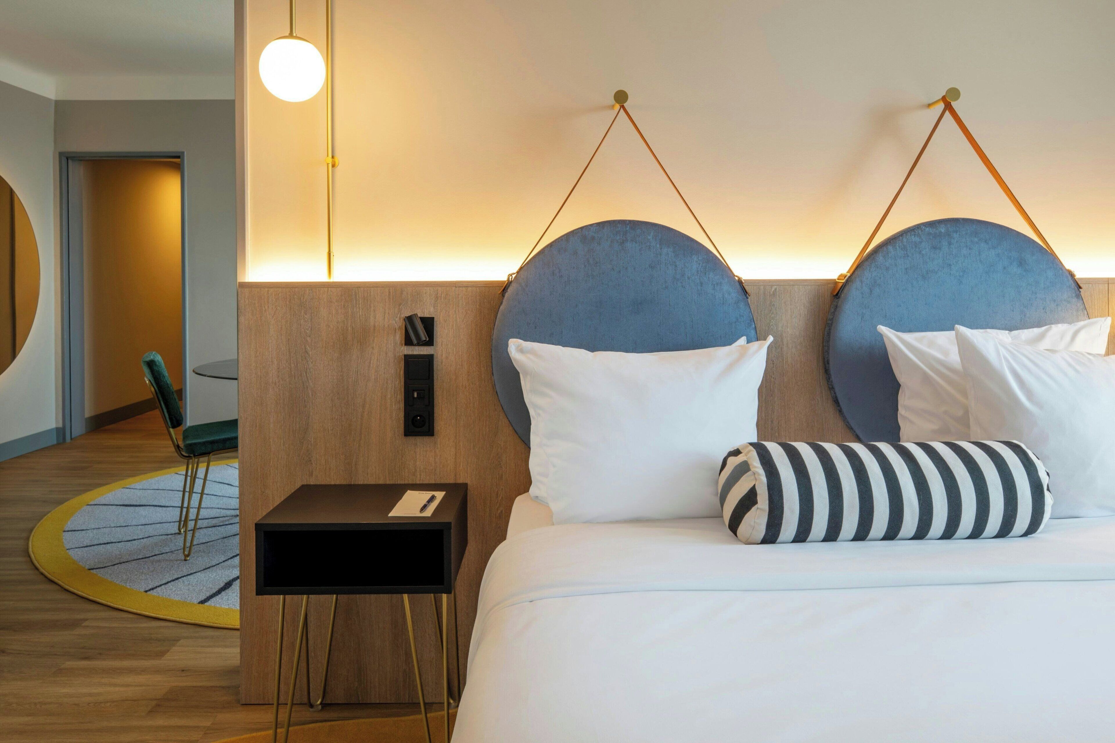 Habitación de hotel con cama, mesita de noche, lámpara y una alfombra redonda.