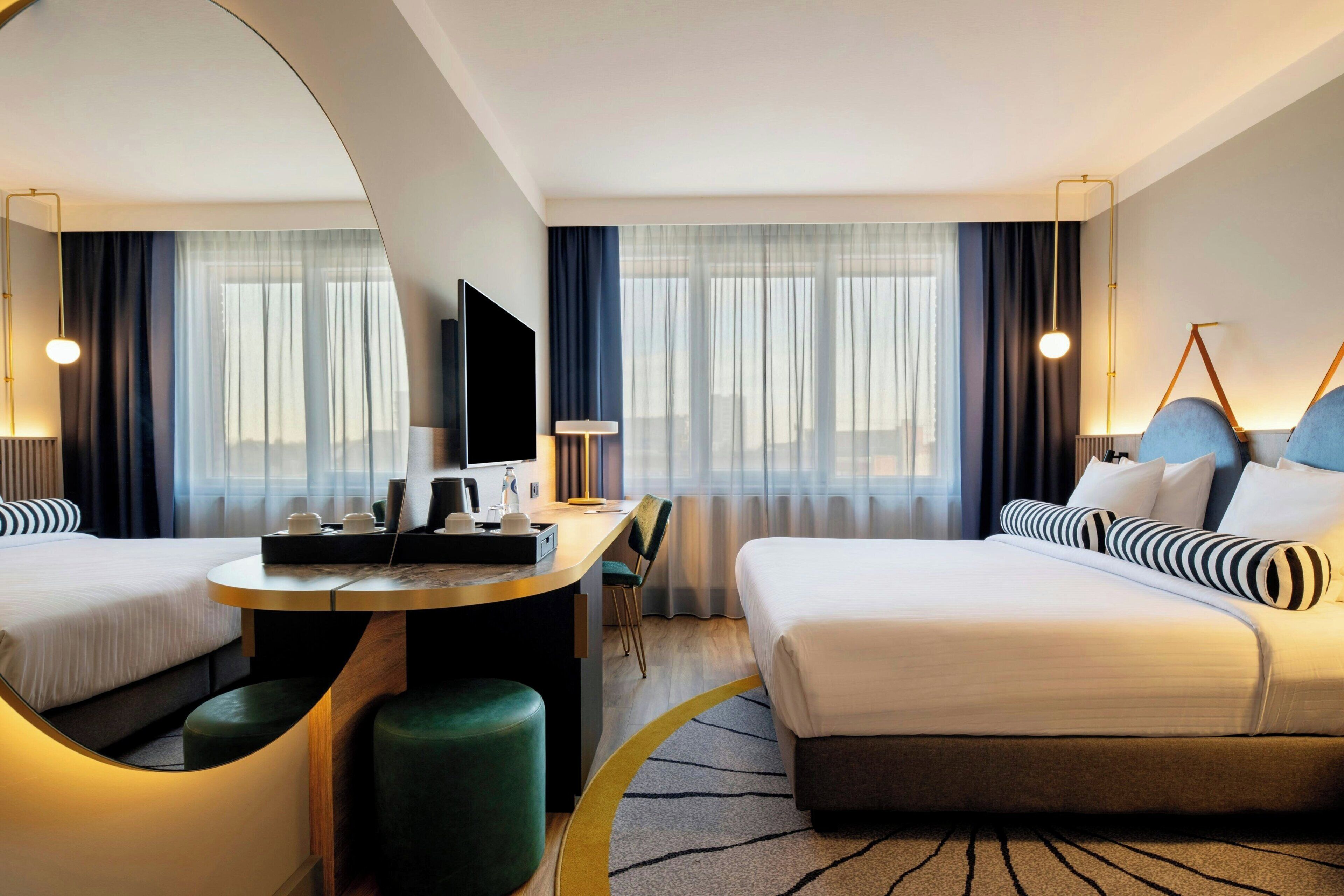 A hotel room with two beds, a desk, a chair, a TV, and a large mirror.