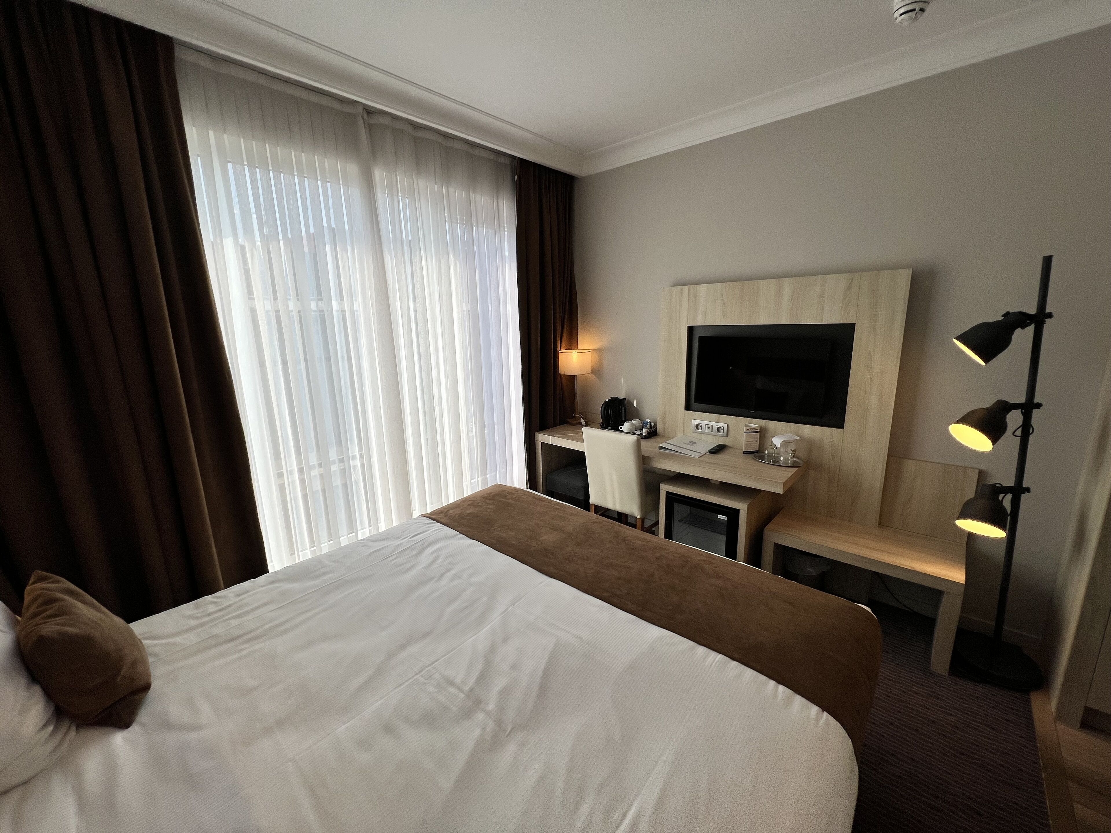 Habitación de hotel con cama, escritorio con televisor, lámpara y ventana con cortinas.