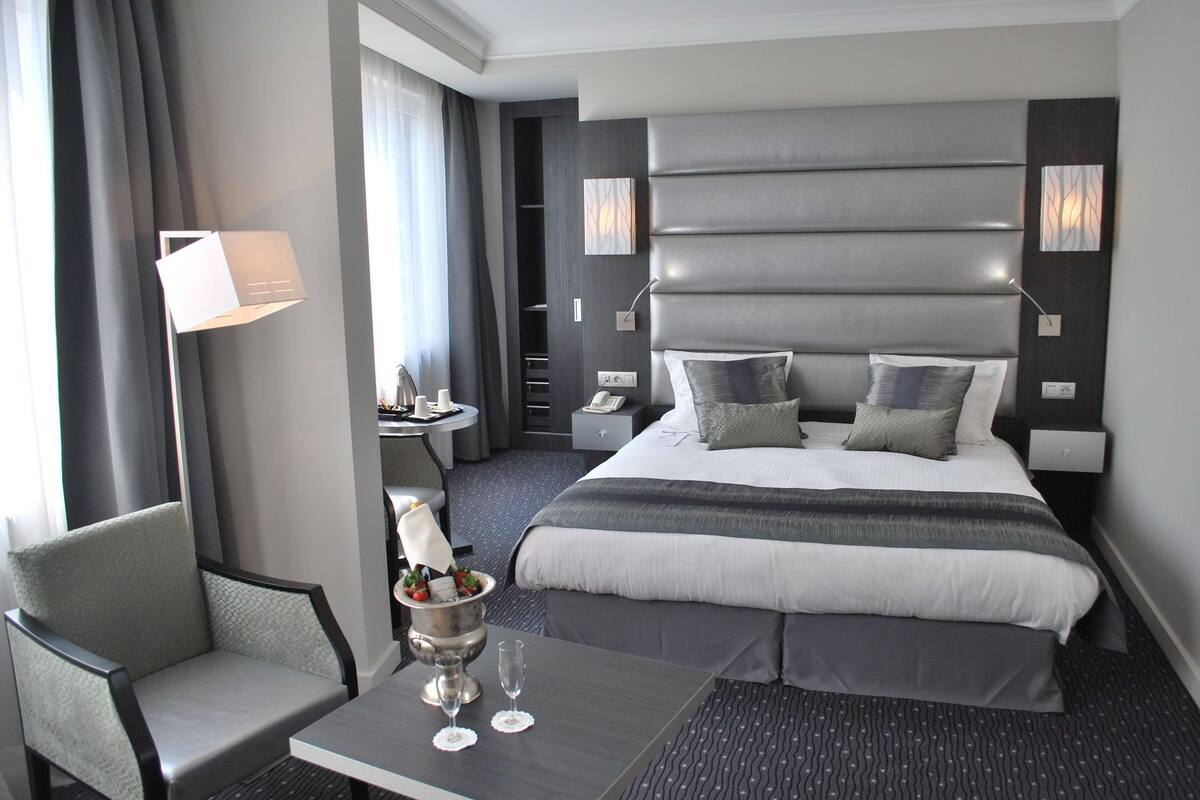 Una habitación de hotel moderna con una cama grande, una mesita de noche con lámpara, una silla y una pequeña mesa con un florero y dos vasos.