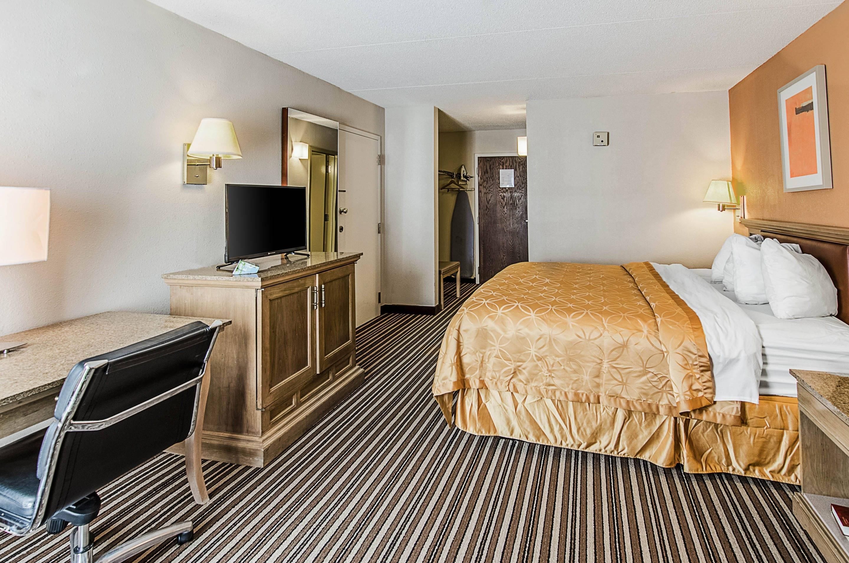 A hotel room with a bed, a desk with a chair, a television, a wardrobe, and a mirror.