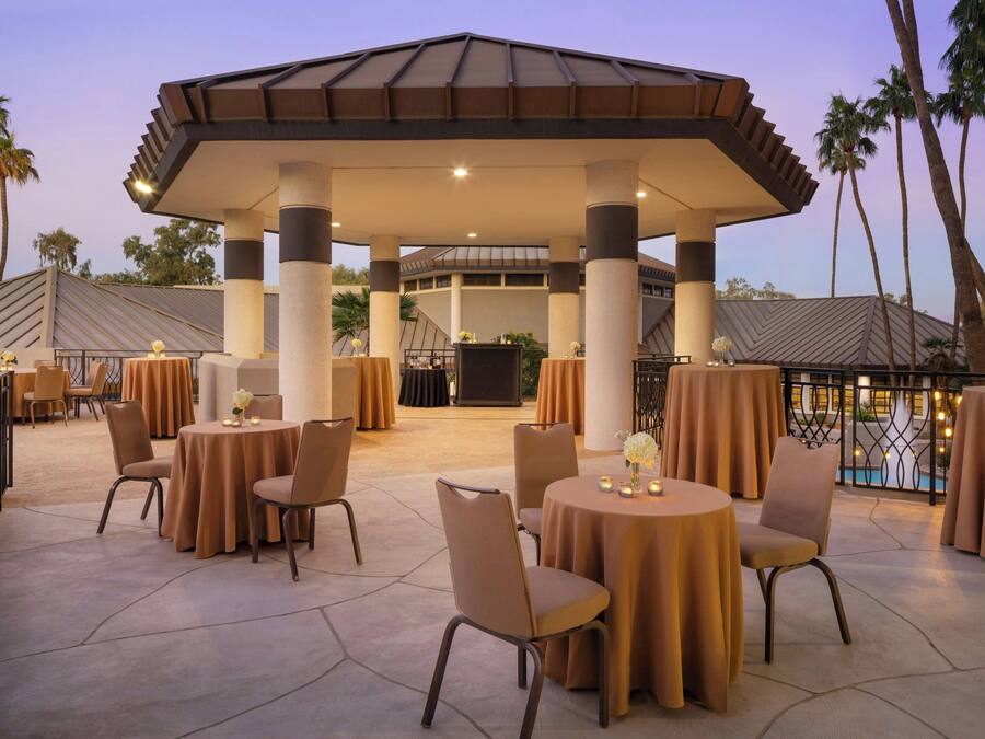 An outdoor patio with round tables and chairs, a covered seating area, and a pool in the background.