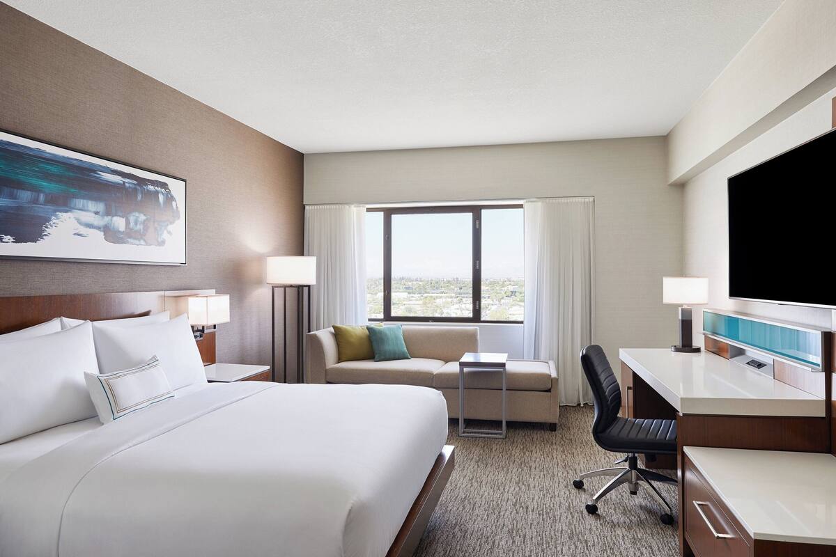 A hotel room with a bed, a sofa, a desk, a chair, a TV, and a window with curtains.