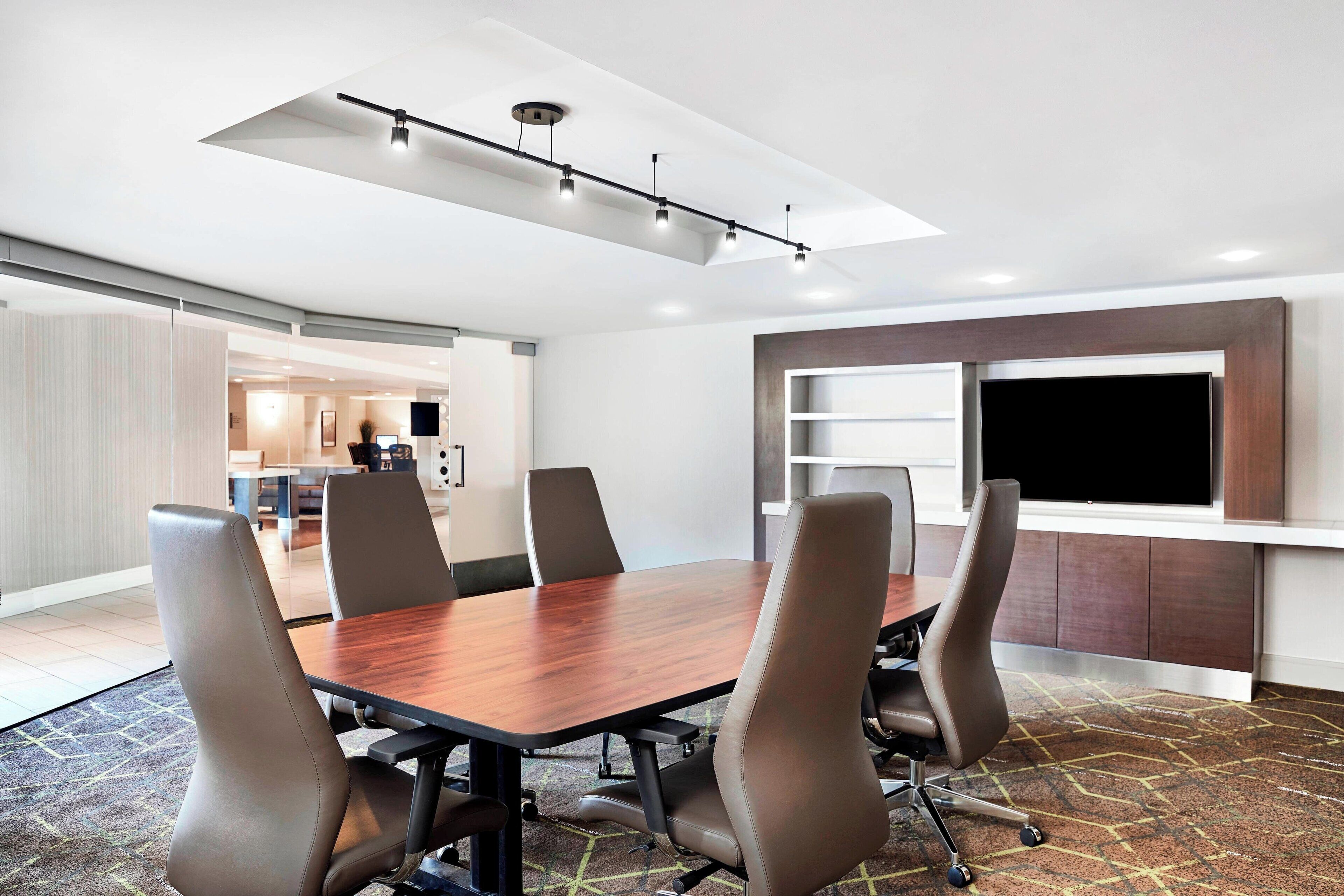 A conference room with a long table, six chairs, a flat-screen TV, and a ceiling track lighting system.