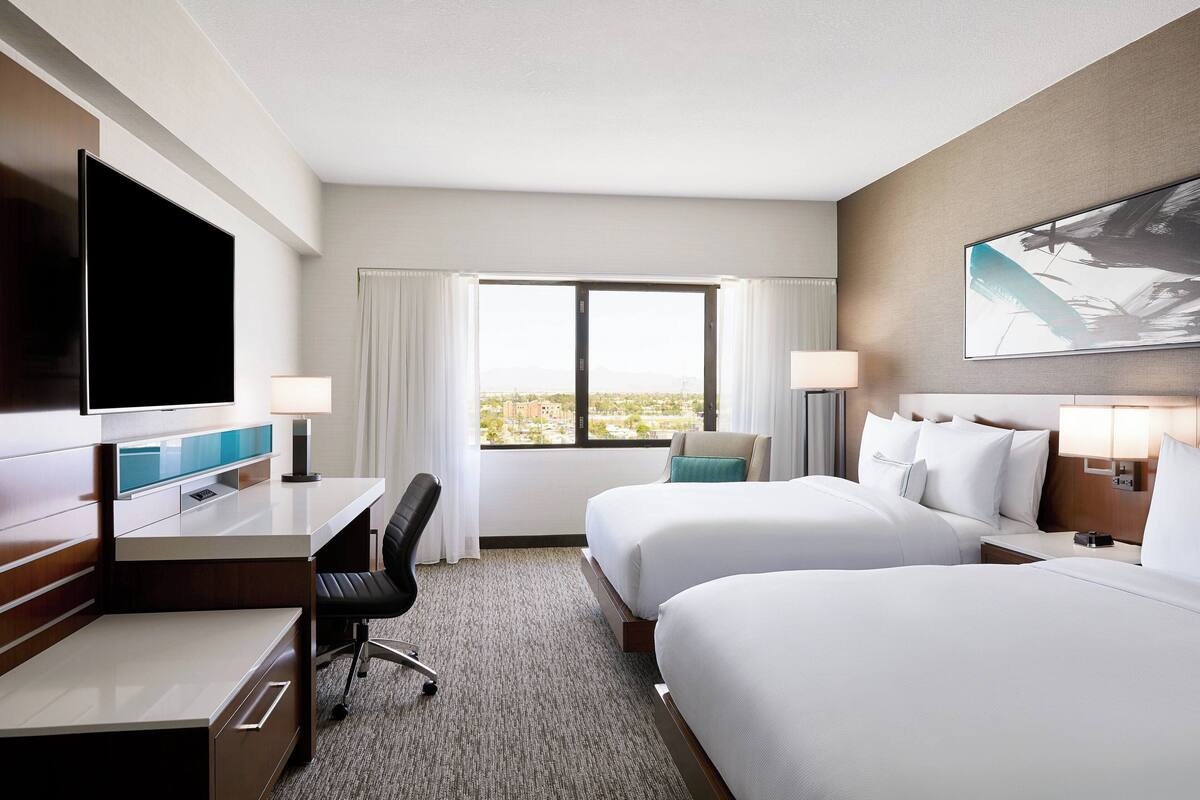 A hotel room with a desk, two beds, a TV, and a large window with a view.