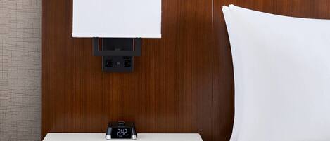 Une chambre d’hôtel avec une tête de lit en bois, une table de chevet dotée d’une horloge numérique, un oreiller blanc et, au mur, un tableau représentant une scène de plage.