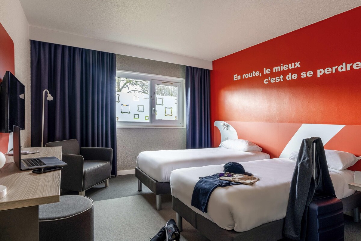 A hotel room with two beds, a desk, a chair, a window with curtains, and a red wall with text.