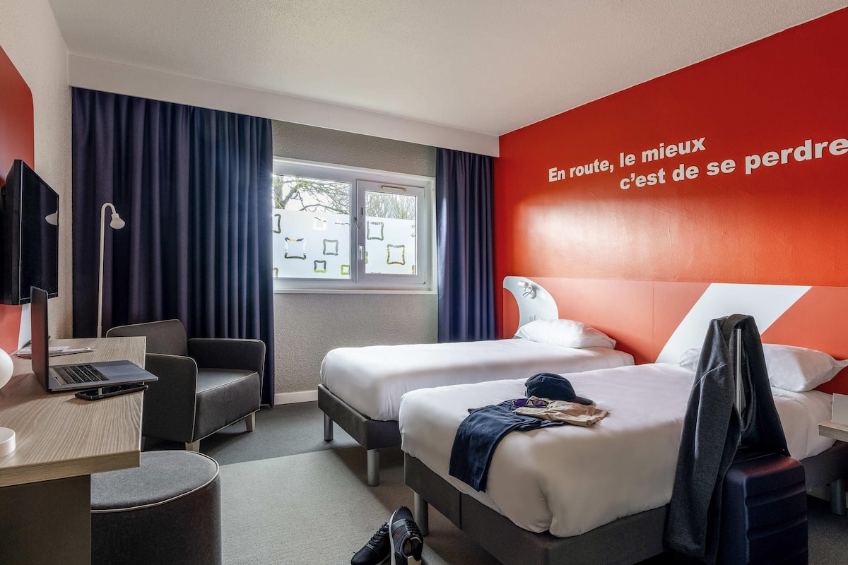 A hotel room with two beds, a desk, a chair, a window with curtains, and a red wall with text.