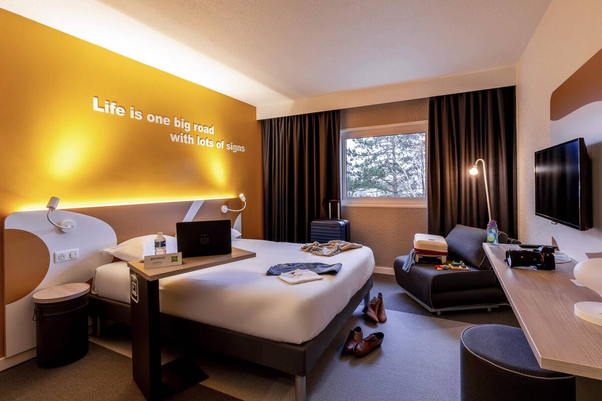 A modern hotel room with a bed, desk, and seating area. A motivational quote is displayed on the wall.
