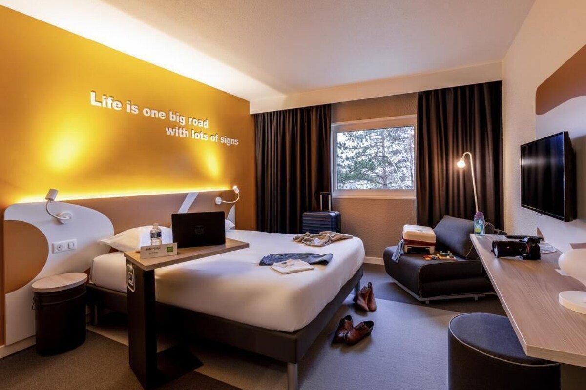 A modern hotel room with a large bed, a desk, a TV, and a wall with motivational text.