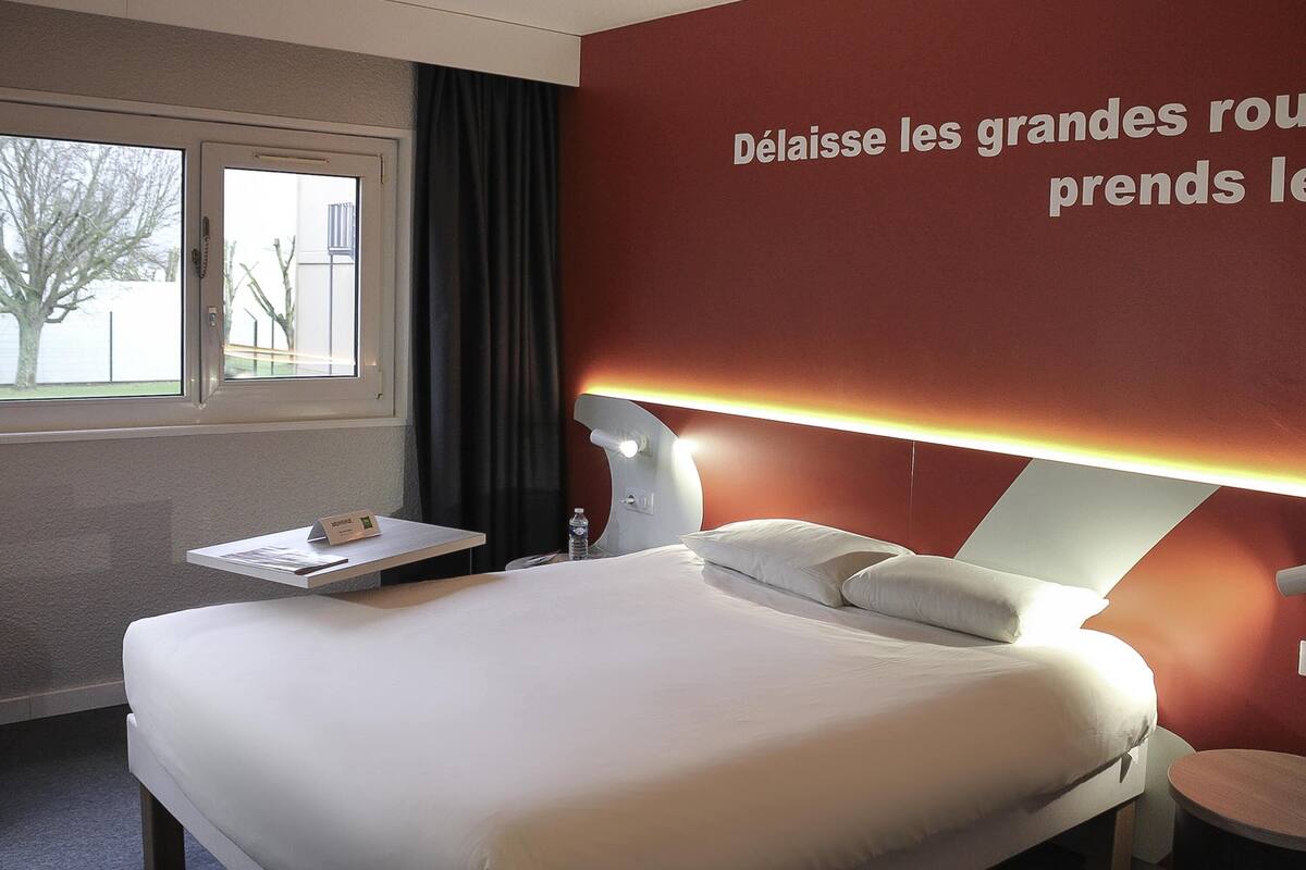 A hotel room with a bed, a bedside table, a lamp, a wall with text, and a window.