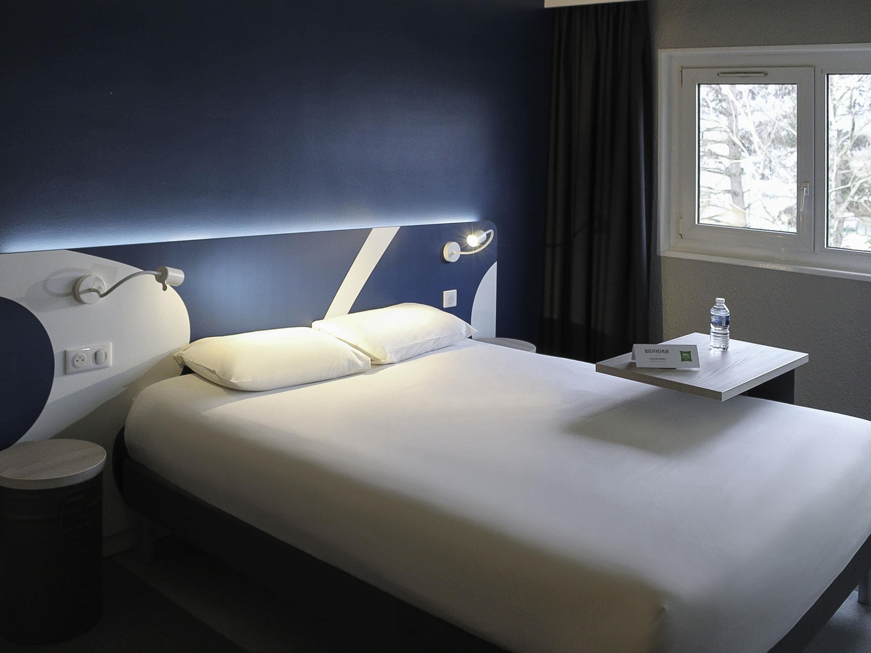 Une chambre d’hôtel avec un lit, une table de chevet avec une bouteille d’eau, une commode et une fenêtre donnant sur des arbres.