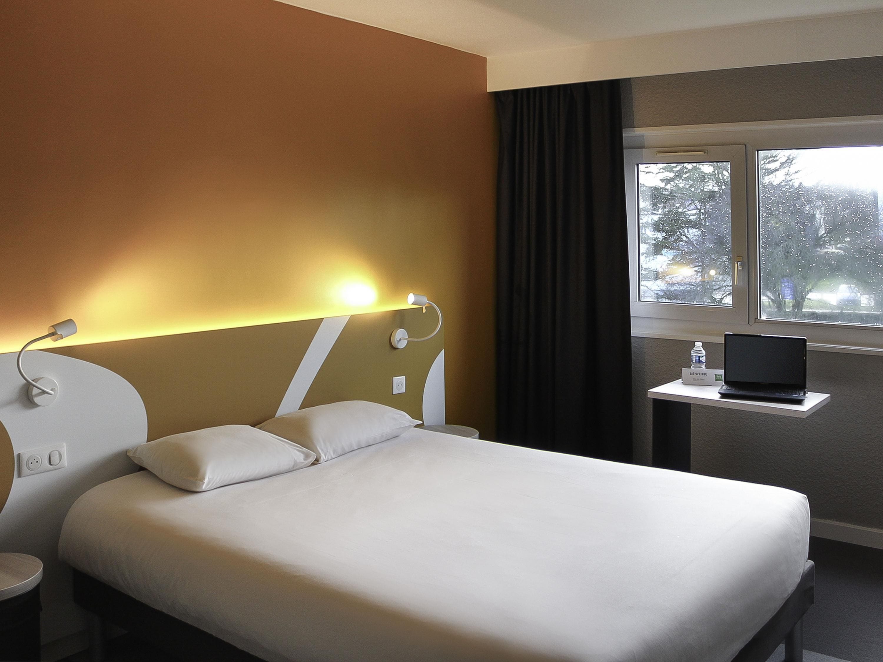 Une chambre d’hôtel avec un lit, un bureau avec un ordinateur portable, une fenêtre donnant sur des arbres et une lampe fixée au mur.