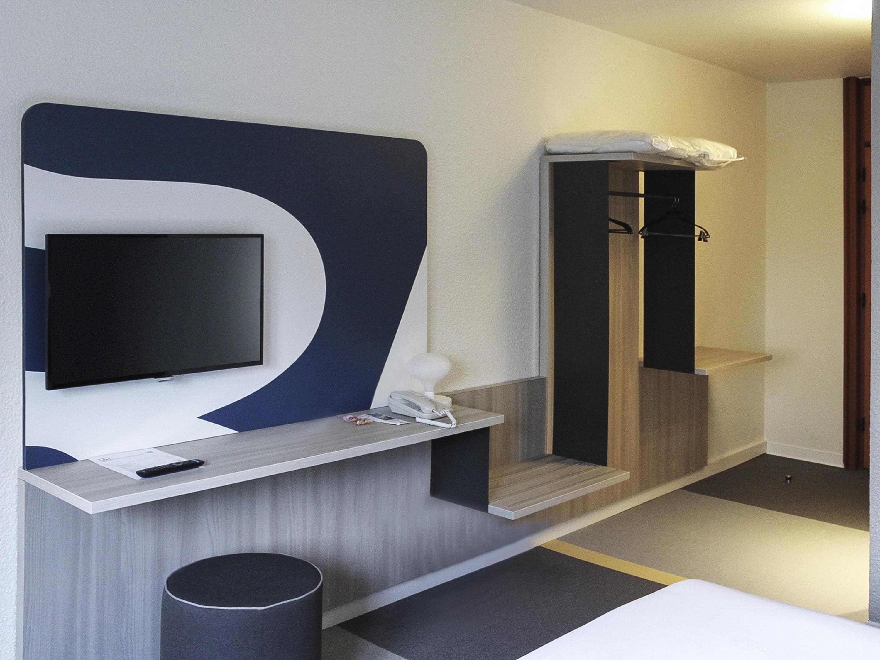 A modern hotel room with a flat-screen TV mounted on a wall, a bedside table with a phone and remote control, a small trash bin, and a shower area with a towel on the wall.