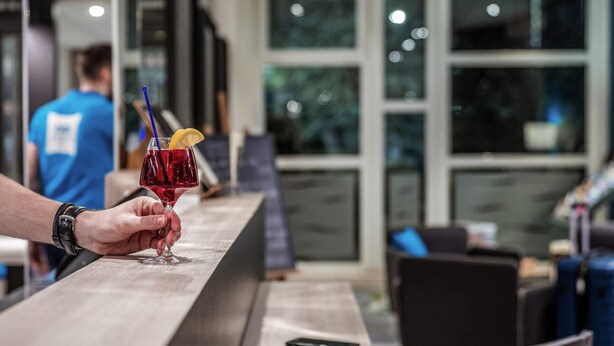 Une main tenant un verre à cocktail avec une tranche de citron et une paille, posé sur le comptoir en bois d’un bar situé dans le hall d’un hôtel.