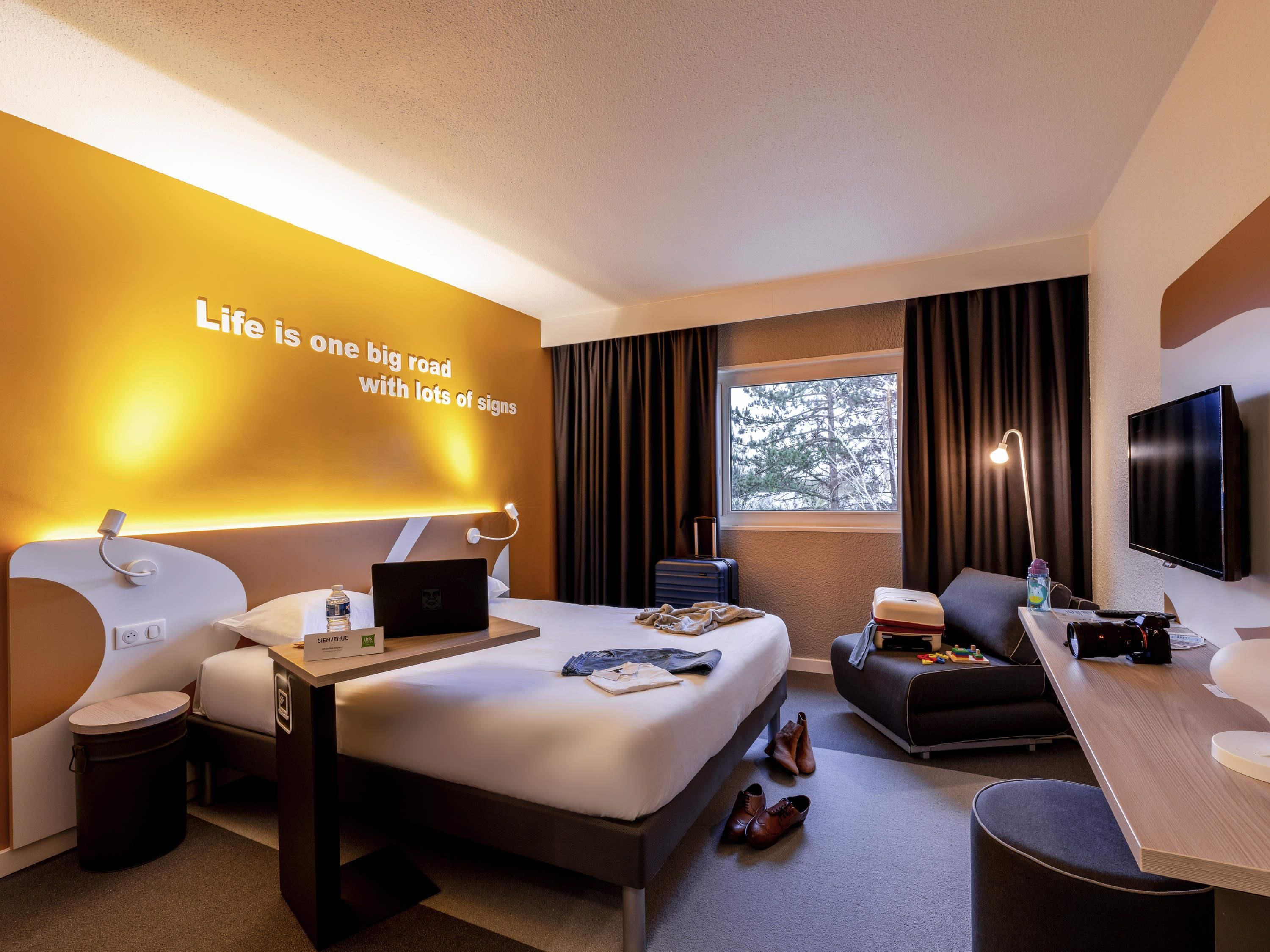 Une chambre d’hôtel moderne, équipée d’un lit, d’un bureau et d’un coin salon. Un proverbe inspirant est affiché sur le mur.