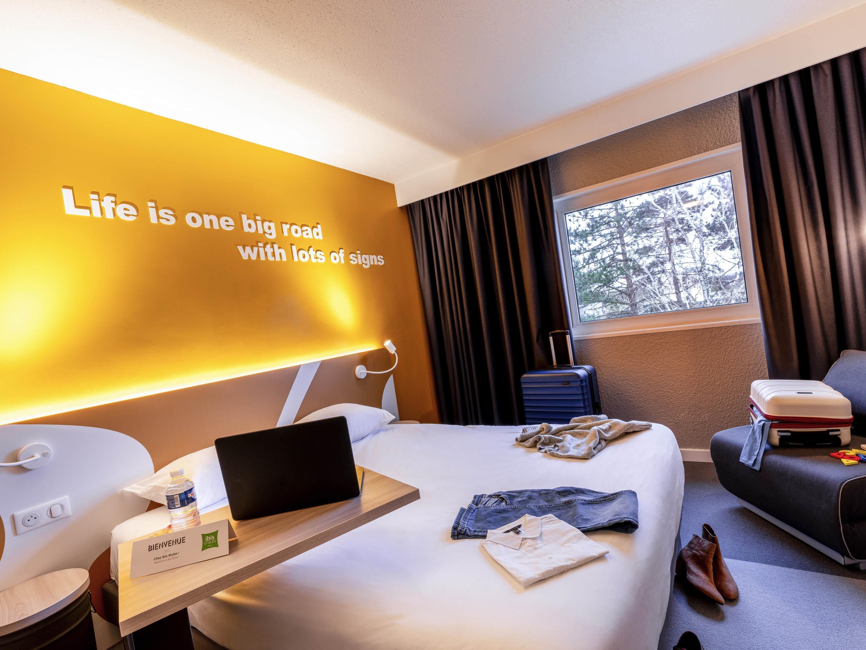 Habitación de hotel con cama, escritorio, computadora portátil, maleta y pared con texto motivacional.