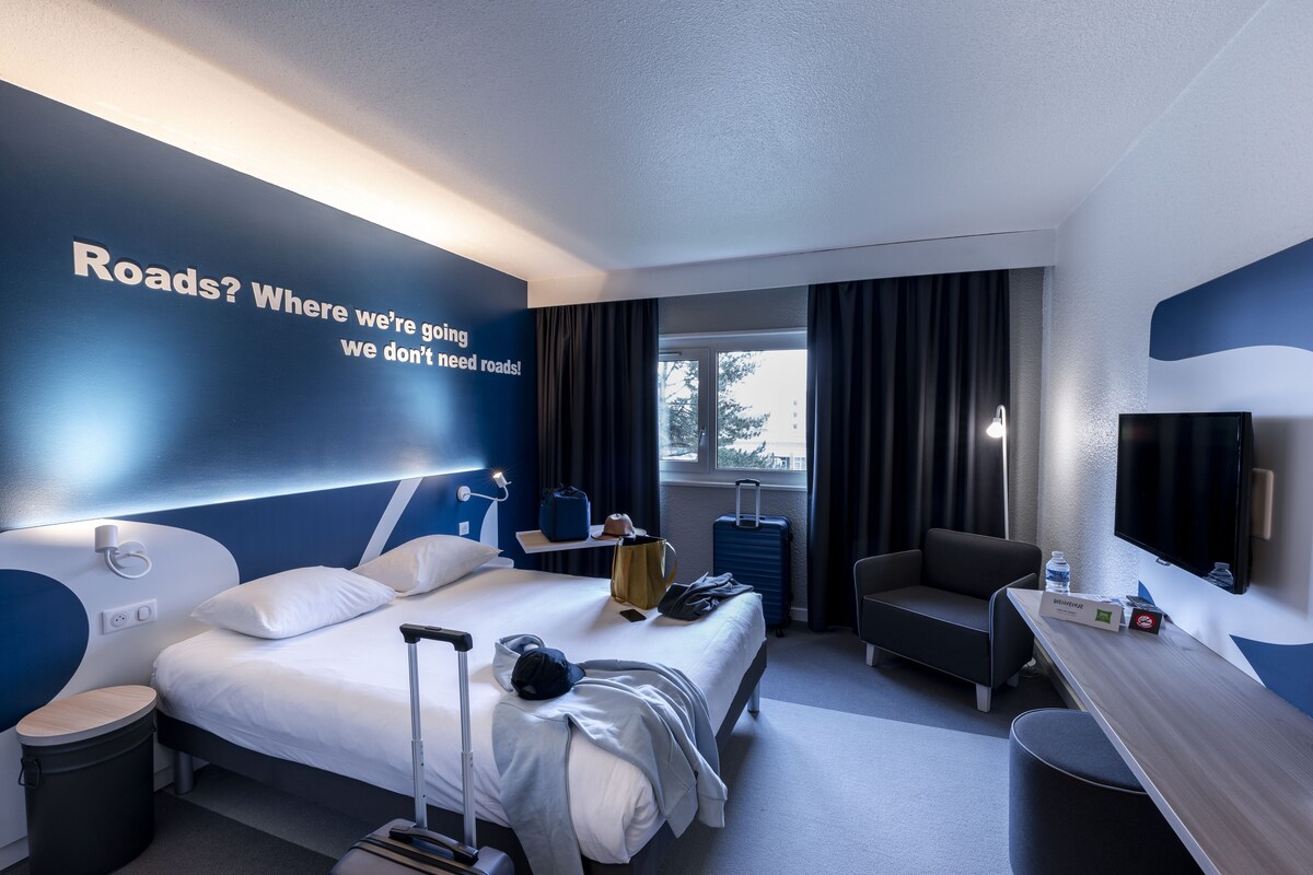 A modern hotel room with a large bed, a desk, a chair, a TV, and a window with curtains.