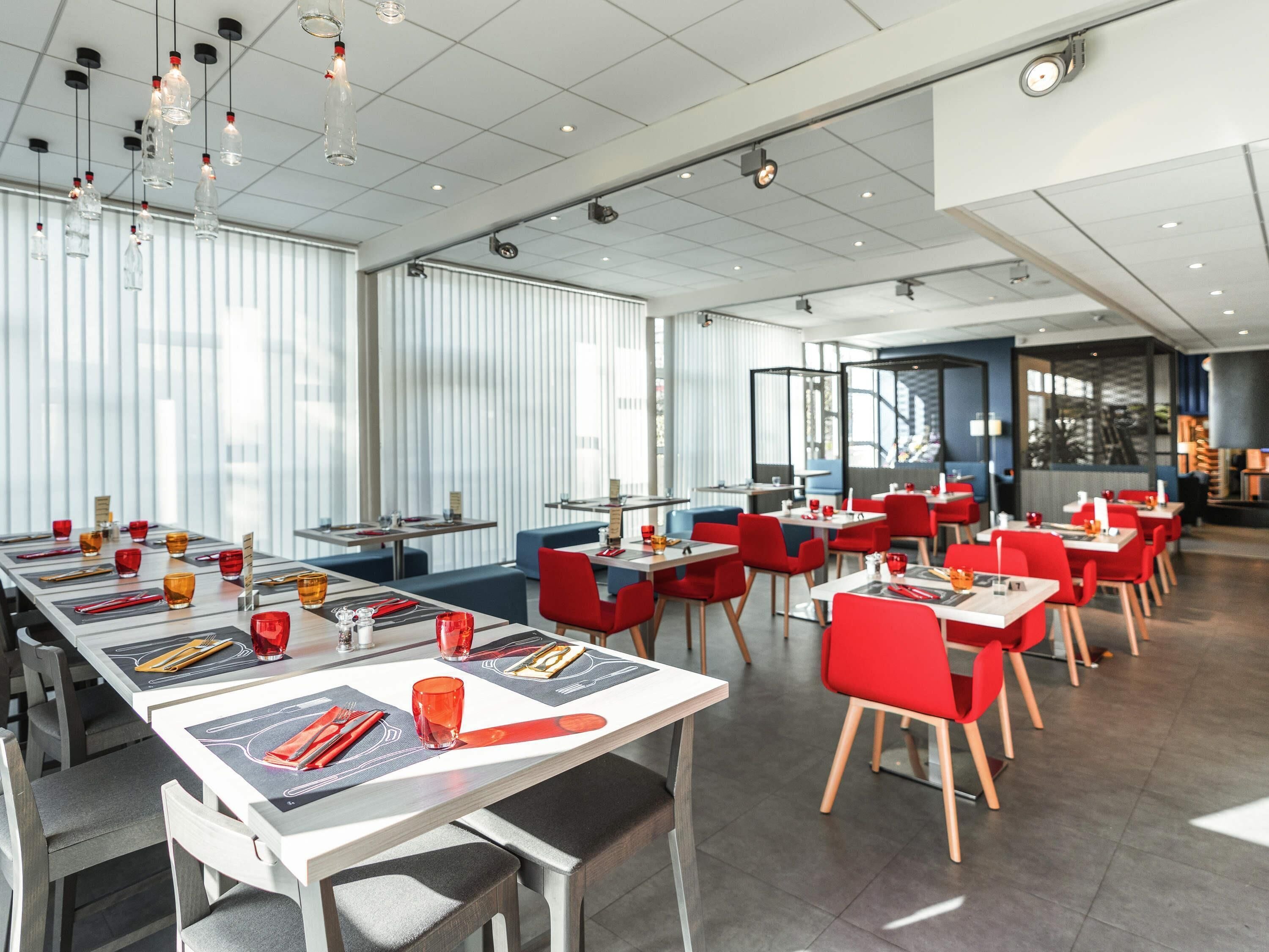 A modern restaurant with long tables, red chairs, and hanging light fixtures.