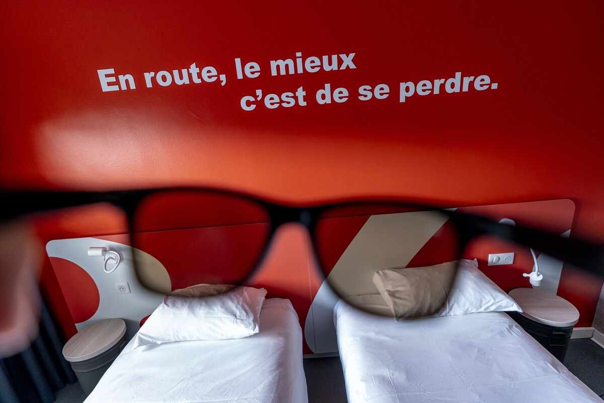 A hotel room with two beds, a red wall with a French quote, and a mirror reflecting the room.