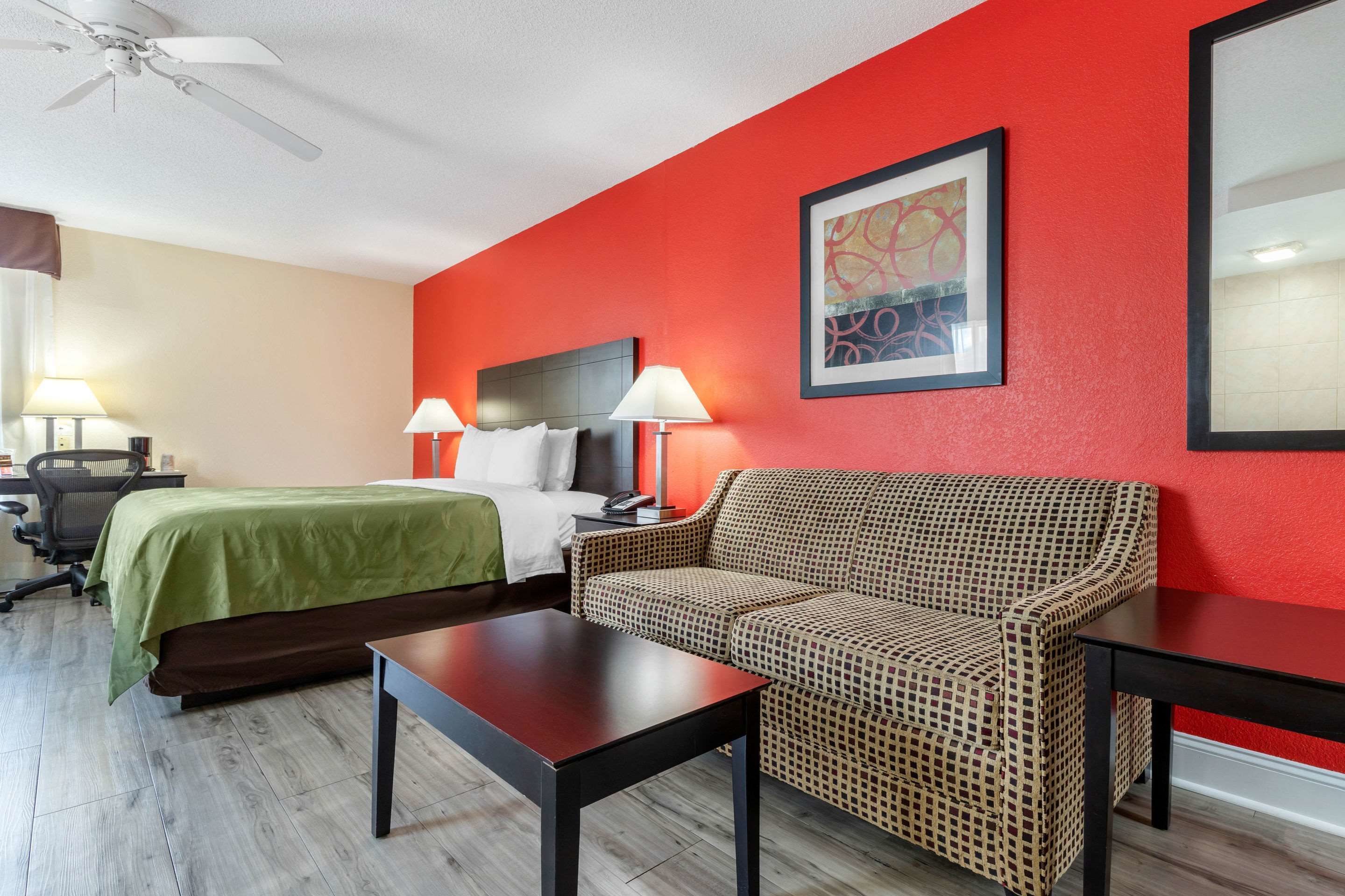 A hotel room with a bed, two bedside tables with lamps, a sofa, a desk with a chair, and a ceiling fan.
