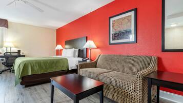 Ein Hotelzimmer mit einem Bett, zwei Nachttischen mit Lampen, einem Sofa, einem Schreibtisch mit Stuhl und einem Deckenventilator.