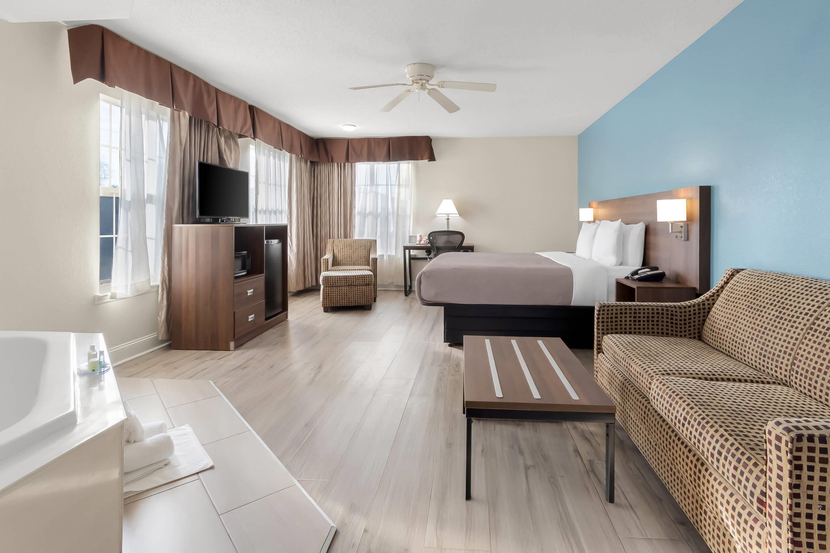 A hotel room with a bed, a sofa, a TV, a desk, a chair, a lamp, a ceiling fan, and a bathroom with a bathtub and toilet.