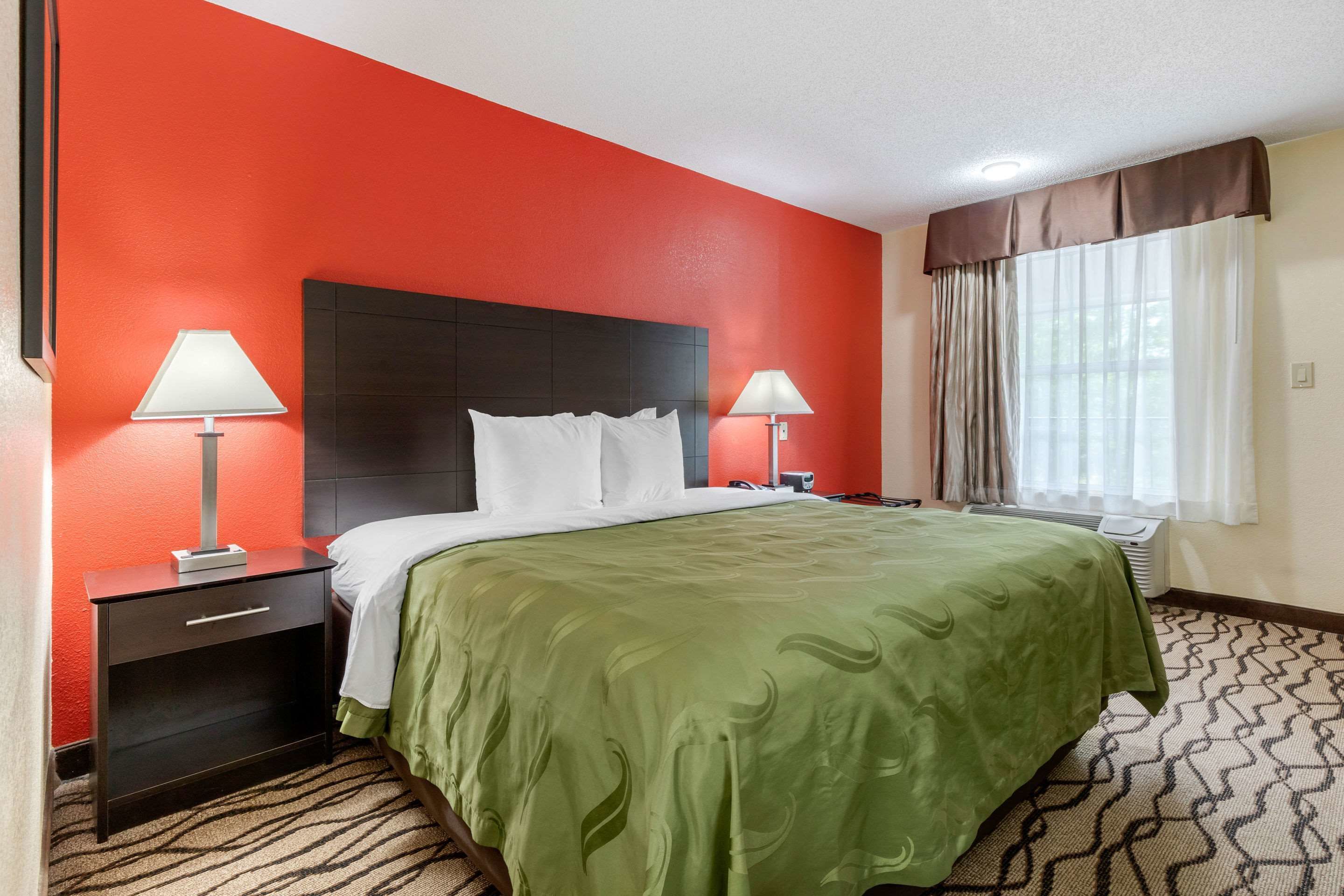 A hotel room with a red accent wall, a large bed with a green bedspread, two bedside tables with lamps, a window with curtains, and a patterned carpet.
