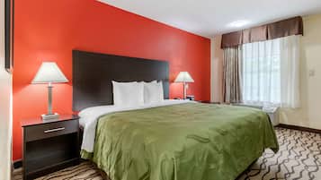 A hotel room with a red accent wall, a large bed with a green bedspread, two bedside tables with lamps, a window with curtains, and a patterned carpet.