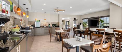 A modern kitchen with a central island, dining area with tables and chairs, and a large flat-screen TV.