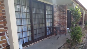 Un patio con pared de ladrillo, grandes puertas correderas de vidrio, una pequeña mesa con sillas y una silla de plástico blanca.