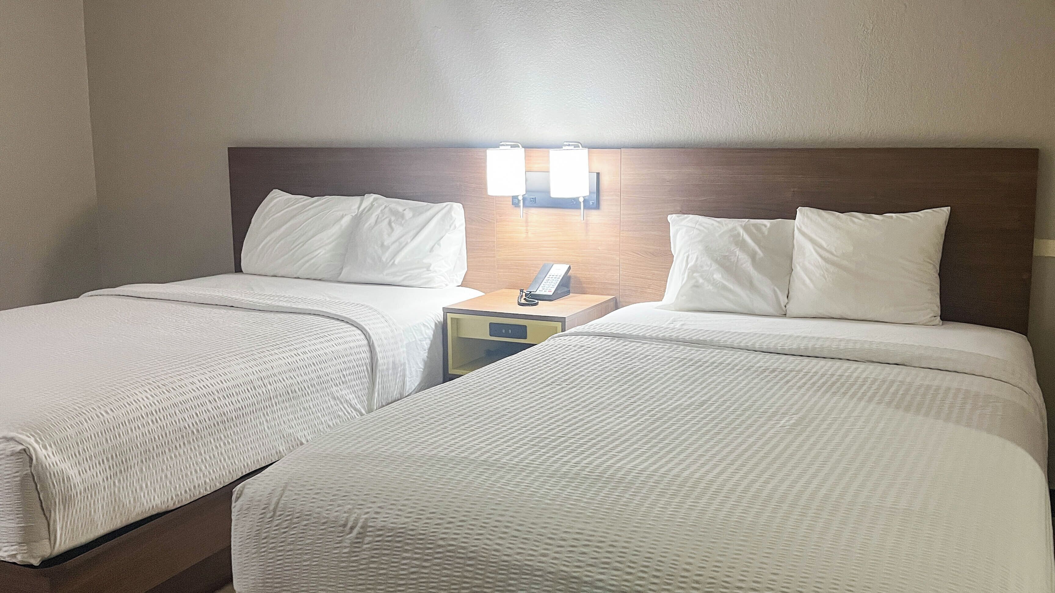 Una habitación de hotel con dos camas, una mesita de noche con teléfono e iluminación en la pared.