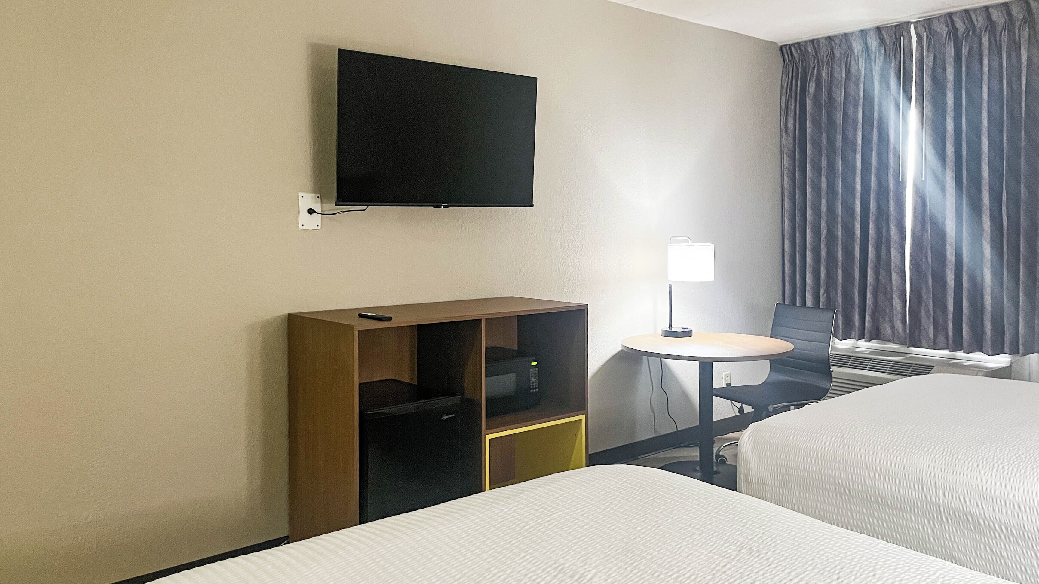 A hotel room with a bed, a desk with a lamp, a television mounted on the wall, and a window with curtains.