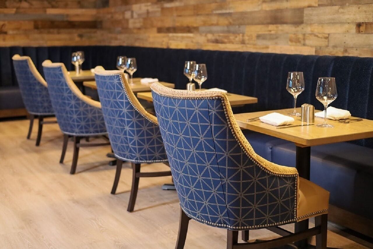 Una fila de sillas tapizadas en azul con diseños geométricos, mesas de madera y un banco acolchado en azul oscuro.