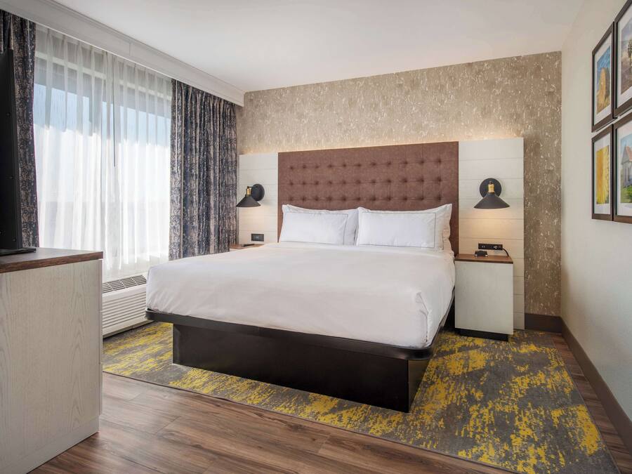 Een hotelkamer met een groot bed, twee bedlampen, een flatscreen televisie, een bureau en een groot raam met dunne gordijnen.