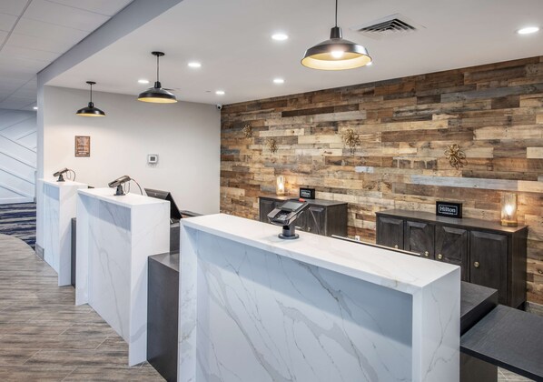 Zona de recepción de hotel con mostradores de mármol, pared de madera e iluminación colgante.