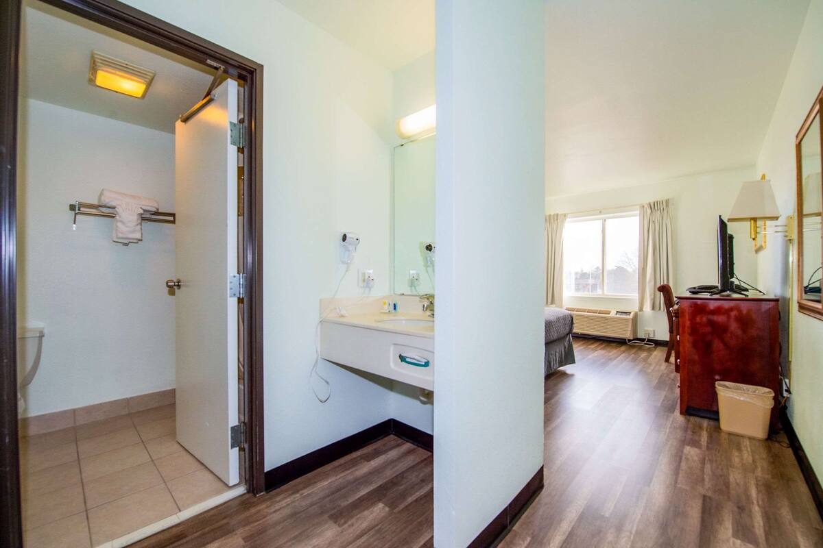 A hotel room with a wooden floor, a bathroom with a toilet, a sink, and a mirror. There is a window with curtains and a lamp on a table.