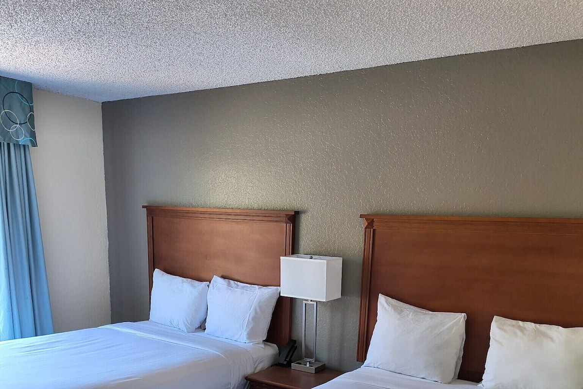 A hotel room with two beds, a nightstand with a lamp, and a window with curtains.