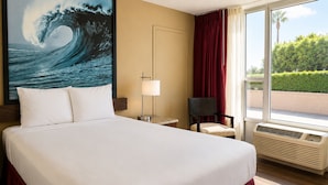 A hotel room with a large bed, a desk, a chair, a window with a view of greenery, and an air conditioning unit. - Kings Inn Anaheim at the Park & Convention Center (Anaheim)