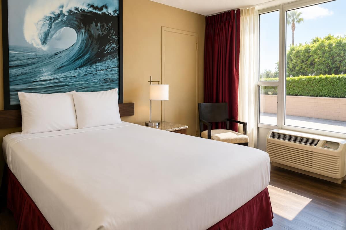 A hotel room with a large bed, a desk, a chair, a window with a view of greenery, and an air conditioning unit.