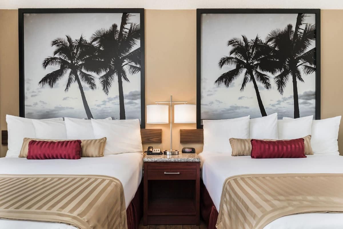 A hotel room with two beds, a nightstand with a lamp, and a large framed picture of palm trees.