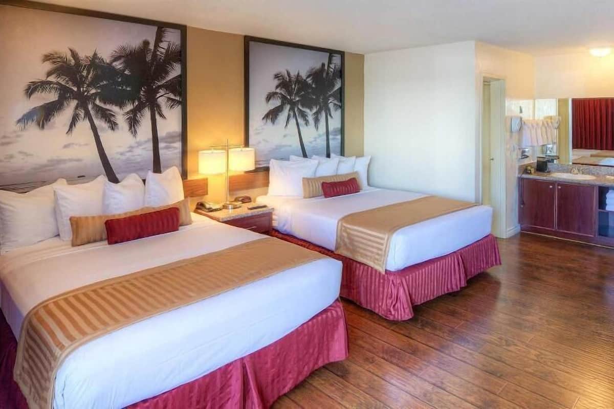 A hotel room with two beds, a nightstand, a lamp, a painting of palm trees, and a view of the ocean.